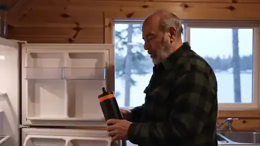 Cabin kitchen at dusk, where a bearded man lifts a tall dark bottle from the refrigerator and shakes it